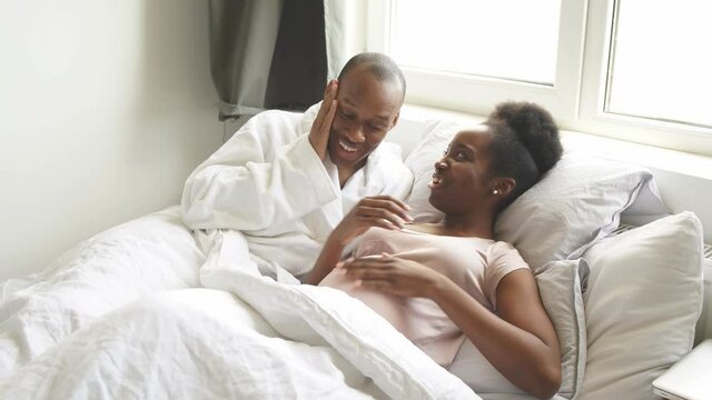 Young black husband cuddling pregnant wife on bed, touch her belly. Happy relationships, pregnancy concept.