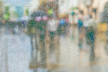 Rain on the window and bokeh city, blurred silhouettes of people, modern urban street. Seasons, weather concept. Selective focus on droplet