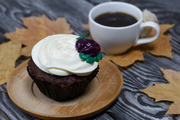 Chocolate muffin with cream flower. A cup of black coffee. Among the dried maple leaves. They lie on pine boards painted in black and white.