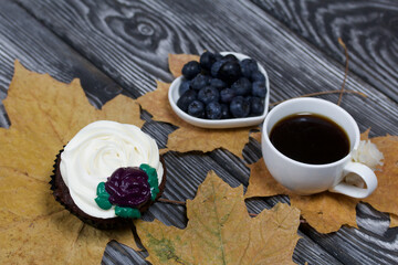 Chocolate muffins with cream flower. A cup of black coffee. Saucer with blueberries. Among the dried maple leaves. Lies on pine boards painted black and white.