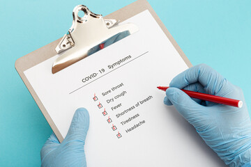 Hands wearing protectives gloves holding a Checklist On Clipboard with COVID-19 symptoms