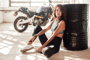 Plakat Sports woman with her custom sport bike on the background resting after riding in white sunny garage.