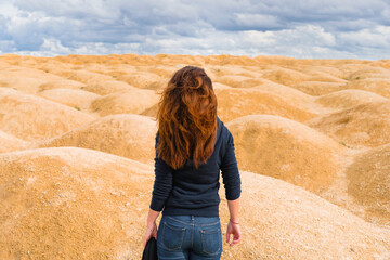 Naklejka premium A young woman with red hair stands with her back to the camera in a desert with sand dunes of bizarre shape