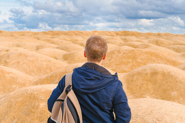 Sporty blond man with a backpack stands with his back to the camera in the desert with sand dunes of bizarre shape