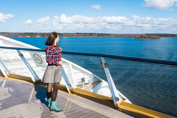 Fototapeta premium Beautiful slender girl traveler on the upper deck of cruise ship. Asian young woman dressed in red plaid shirt and short skirt in luxury cruise. Elegant happy woman look at ocean relax on luxury liner