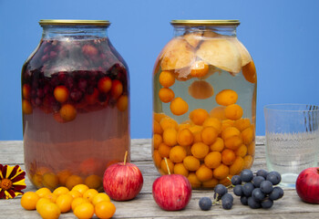 Two-litre cans with compote stand on a wooden surface. There's a blue wall in the back. Homemade harvests from plums, apples, grapes