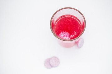 In a glass with pink water, a soluble, instant or dissolve pink tablet bubbles. Nearby are similar tablets: vitamin C, dietary supplements, painkillers. White background, copy space