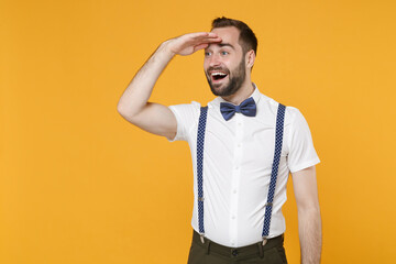 Excited young bearded man 20s in white shirt bow-tie suspender posing standing holding hand at forehead looking far away distance isolated on bright yellow color wall background studio portrait.