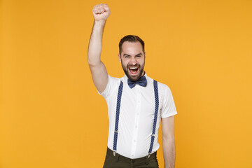 Cheerful screaming young bearded man 20s in white shirt bow-tie suspender posing standing standing rising hand up clenching fist isolated on bright yellow color wall background studio portrait.