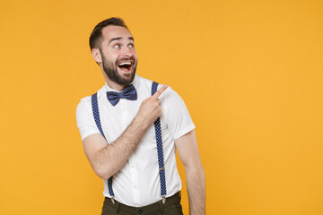 Excited young bearded man 20s in white shirt bow-tie suspender posing standing pointing index finger aside up on mock up copy space isolated on bright yellow color wall background studio portrait.