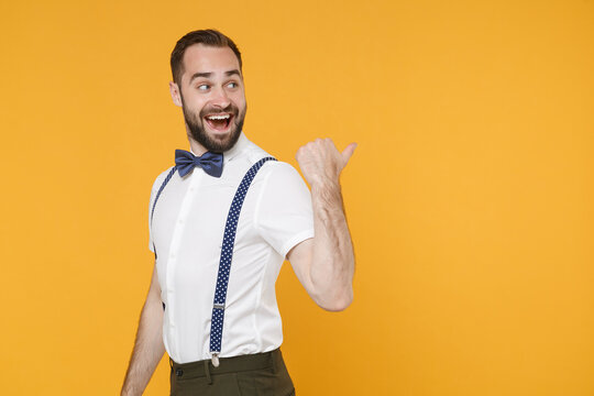 Side view of funny young bearded man 20s wearing white shirt bow-tie suspender posing standing pointing thumb aside on mock up copy space isolated on bright yellow color background studio portrait.