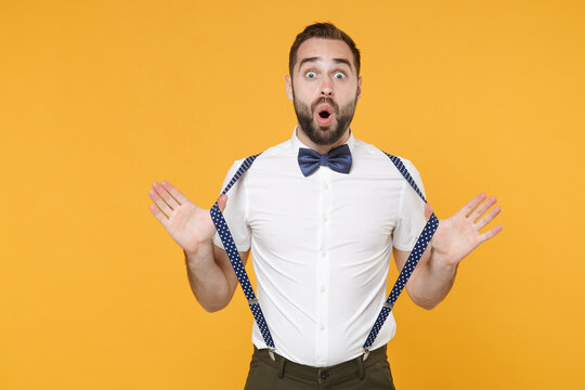 Shocked amazed young bearded man 20s wearing white shirt bow-tie posing standing stretching suspender spreading hands looking camera isolated on bright yellow color wall background studio portrait.