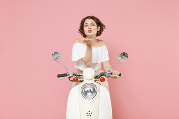 Charming pretty young brunette woman 20s wearing white summer clothes blowing sending air kiss making duck face looking camera driving moped isolated on pastel pink colour background, studio portrait.