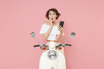 Shocked young brunette woman 20s wearing white summer clothes using mobile cell phone typing sms message put hand on cheek sit driving moped isolated on pastel pink colour background studio portrait.