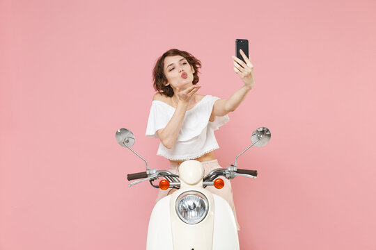 Pretty young brunette woman 20s wearing white summer clothes posing doing selfie shot on mobile phone blowing sending air kiss driving moped isolated on pastel pink colour background, studio portrait.