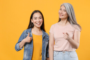 Smiling funny family two asian women girls gray-haired mother and brunette daughter in casual clothes posing pointing index finger on camera isolated on yellow color wall background studio portrait.
