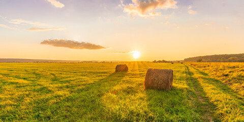 Scenic view at picturesque burning sunset in a green shiny field with hay stacks, bright cloudy sky , golden sun rays and road leading far away, summer valley landscape