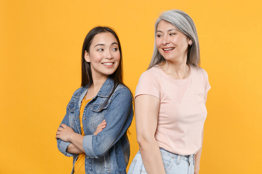 Smiling Family Asian Women Girls Gray-haired Mother And Brunette Daughter In Casual Clothes Posing Standing Back To Back Looking At Each Other Isolated On Yellow Color Background Studio Portrait.