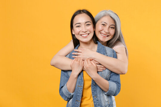 Smiling cheerful pretty family asian female women girls gray-haired mother and brunette daughter in casual clothes posing hugging looking camera isolated on yellow color background studio portrait.