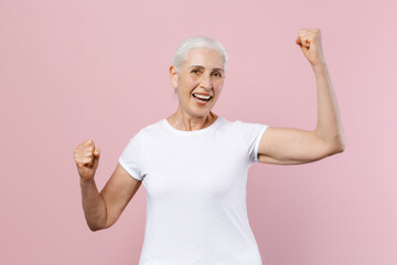 Happy joyful elderly gray-haired female woman wearing white design casual t-shirt posing clenching fists doing winner gesture looking camera isolated on pastel pink color background studio portrait.