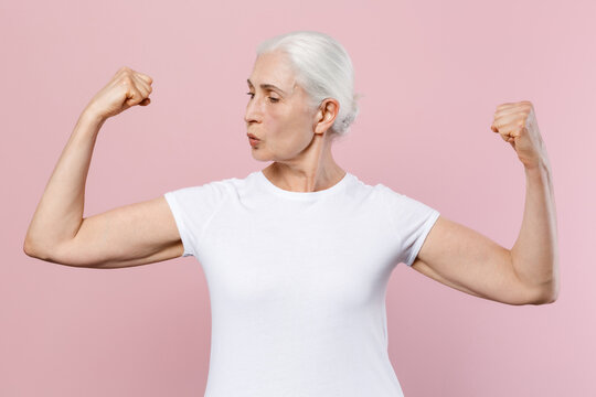 Strong shocked elderly gray-haired female woman 60s 70s wearing white design casual t-shirt posing showing biceps muscles looking aside isolated on pastel pink color wall background studio portrait.