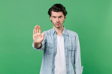 Serious young brunet man 20s wearing casual clothes white t-shirt denim shirt posing standing showing stop gesture with palm looking camera isolated on green color wall background studio portrait. © ViDi Studio