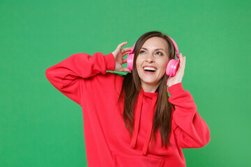 Cheerful young brunette woman 20s wearing bright red casual streetwear hoodie posing listening music with headphones looking up put hands on head isolated on green color background studio portrait.