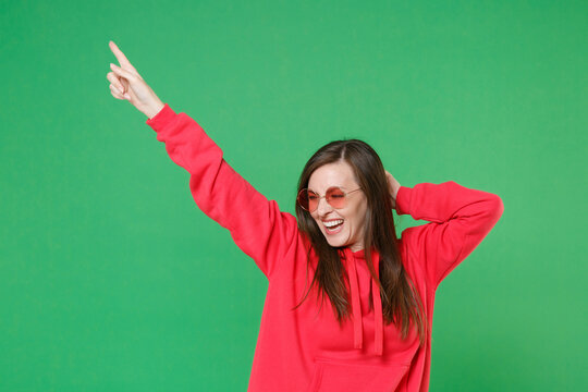 Cheerful laughing young brunette woman 20s in bright red casual streetwear hoodie eyeglasses posing dancing rising hands pointing index finger up isolated on green color background studio portrait.