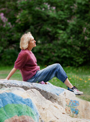 Naklejka na ściany i meble Pretty blond woman sitting on the giant stone ib the park, relaxing after long walk, hard day. Horizontal shot