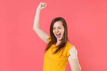 Happy overjoyed screaming young brunette woman 20s in yellow casual t-shirt posing standing clenching fists doing winner gesture keeping eyes closed isolated on pink color background studio portrait.