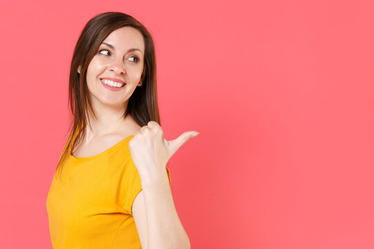 Side view of smiling cheerful attractive young brunette woman 20s wearing yellow casual t-shirt posing pointing thumb aside on mock up copy space isolated on pink color background studio portrait.