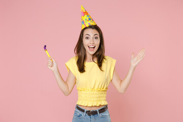 Excited surprised young brunette woman 20s wearing yellow casual t-shirt birthday hat posing standing celebrating holding pipe spreading hands isolated on pastel pink color background studio portrait.