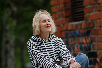 Beautiful blond women wearing casual striped hoodie outdoors, sitting near graffiti-painted brick wall