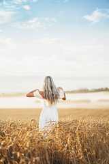 Portrait of a beautiful young blonde woman in a white dress running through the golden wheat field in the down with fog and blue sky. View from the back. Country, nature, summer holidays, agriculture.