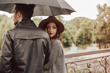 Back view of heterosexual couple standing outside on the bridge in the city park near the river in cloudy rainy moody weather with black umbrella. Image with copy space and bokeh background.