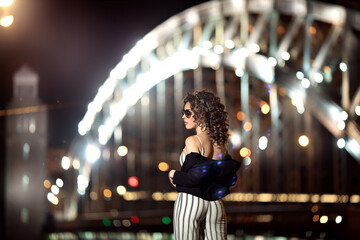 A girl in white clothes with black stripes stands with her back against the background of the bridge. Portrait of a girl in the style of the 90s. Model on the background of the night bridge.