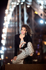 A girl in white clothes with black stripes sits on the background of the bridge. Portrait in the style of the 90s. Girl with curls on the background of the night bridge.