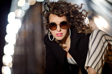 A curly-haired girl in sunglasses and white earrings sits against the bridge. Portrait in the style of the 90s
