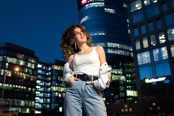 A girl in a white t-shirt and jeans against the background of night houses. Portrait of a girl in the style of the 90s. Model with curls and pink lipstick on the background of an urban landscape.