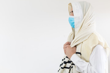 Orthodox Jewish man praying with prayer shawl, tallit, over his head, wearing protective mask...