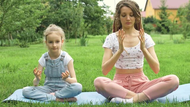 Young mother with dreadlocks and little daughter are doing yoga exercises in lotus position on grass in the park at the day time. Concept of friendly family and of summer vacation.