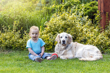 Cute liitle baby boy with open mouth is posing with a dog in the garden.