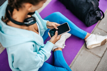 Young adult woman resting after fitness training. She is sitting and using her smart phone to communicate with someone.