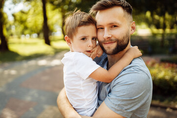 Fototapeta na wymiar Portrait of handsome man with little boy at the park, lovely son sitting on fathers arms, smiling, dad and toddler spend time together, enjoy tender family moments, parenthood concept