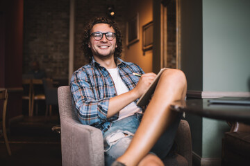 Cheerful curly millennial hipster guy in trendy eyewear sitting in cafe interior with planner making notes on free time, happy creative male designer making sketches and drawings on free time smiling