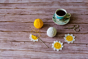 Crocheted daisy, balls of colored yarn, scissors, and a cup of coffee on wooden background
