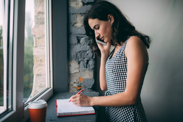 Fototapeta na wymiar Serious caucasian young woman having mobile phone conversation making banking at home interior, pensive millennial female writing in notepad making call via smartphone standing near window sill.