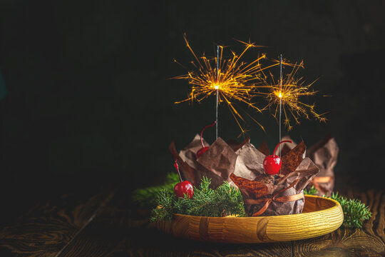 Sweet chocolate muffins with sparklers decorated cherry in brown paper with ribbon on wooden bowl surrounded pine branches. Close up, shallow depth of the field, greeting food composition.