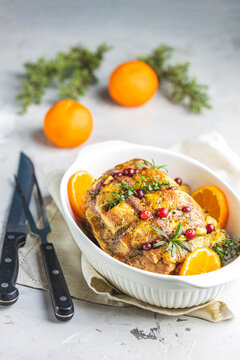 Roasted pork in white dish, christmas baked ham with cranberries, tangerines, thyme, rosemary, garlic on light table surface.