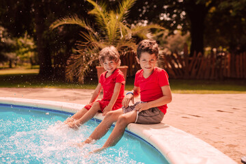 Two beautiful caucasian boys in red T-shirts are sitting on the edge of the pool and spraying water with their feet. Children at the pool by the palm trees